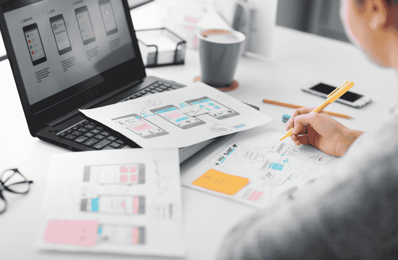 Diseñador creando wireframes de interfaz de usuario para app con IA en portátil y papel.